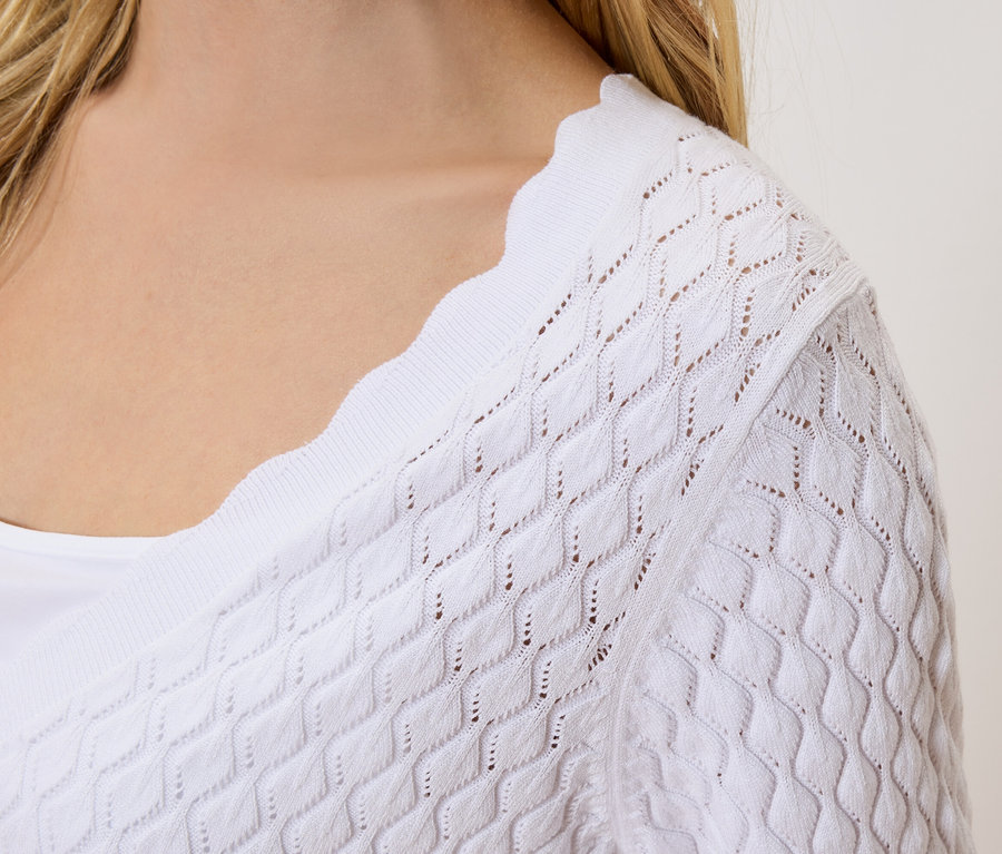 Közeli kép egy szőke hajú nőről, aki egy fehér, áttört kötött kardigánt visel egy fehér felső fölött.