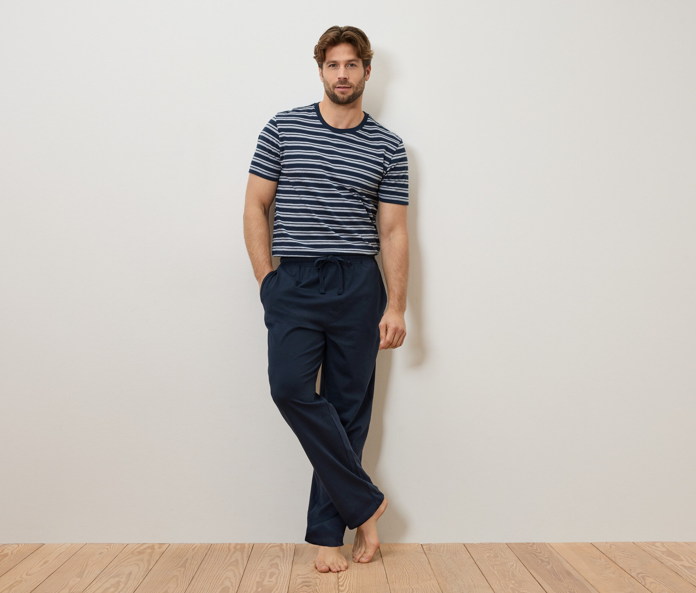 Férfi támaszkodik a falnak, kék-fehér csíkos pólót és sötétkék pizsamaalsót visel.