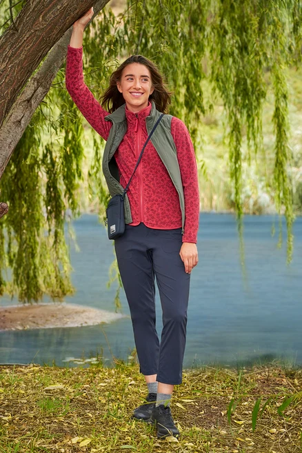 Egy nő egy fa mellett áll a tónál, piros polár pulóvert, sötétkék nadrágot és khaki »packable« Hybrid-Weste mellényt visel.