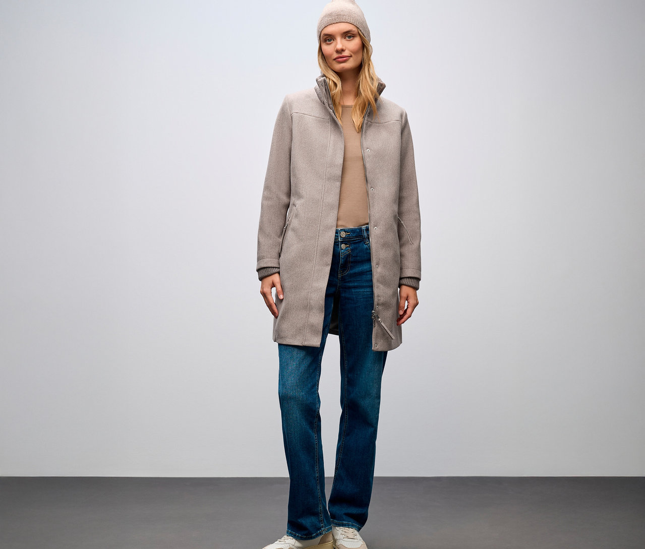 Nő áll, Street One rövid kabátot, farmert és sapkát visel.