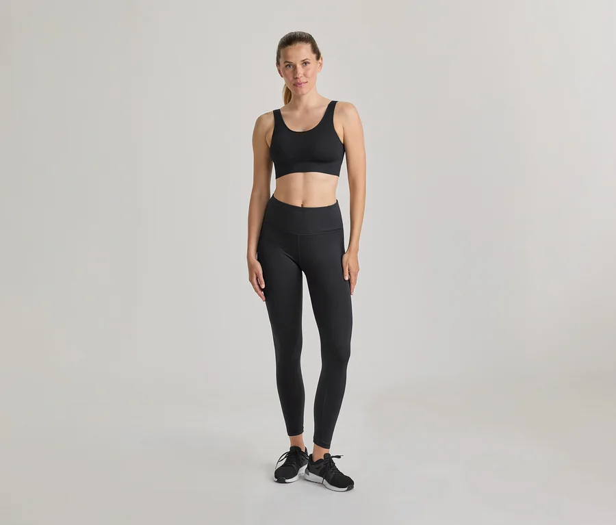 Egy nő áll, fekete sportmelltartót és fekete leggingset visel. Fekete-fehér sportcipő, haja hátrakötve. Világos, egyszínű háttér.