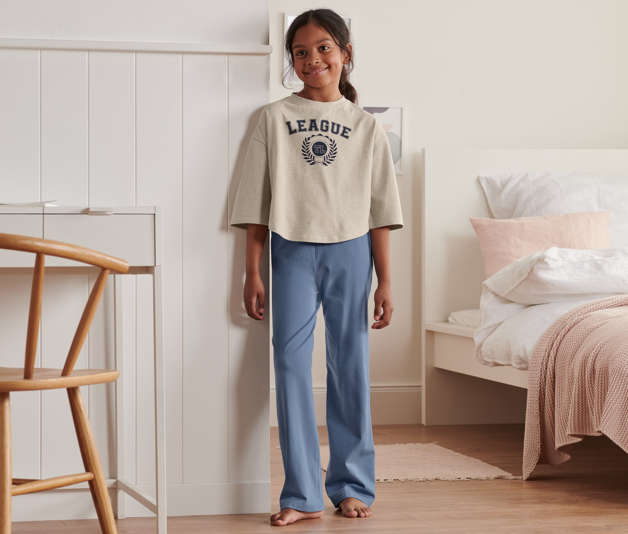 Egy lány áll a hálószobában, és egy bézs pólót és egy kék pizsama nadrágot visel a 2 lány pizsama szettben, bézs/világoskék/sötétkék szettből.