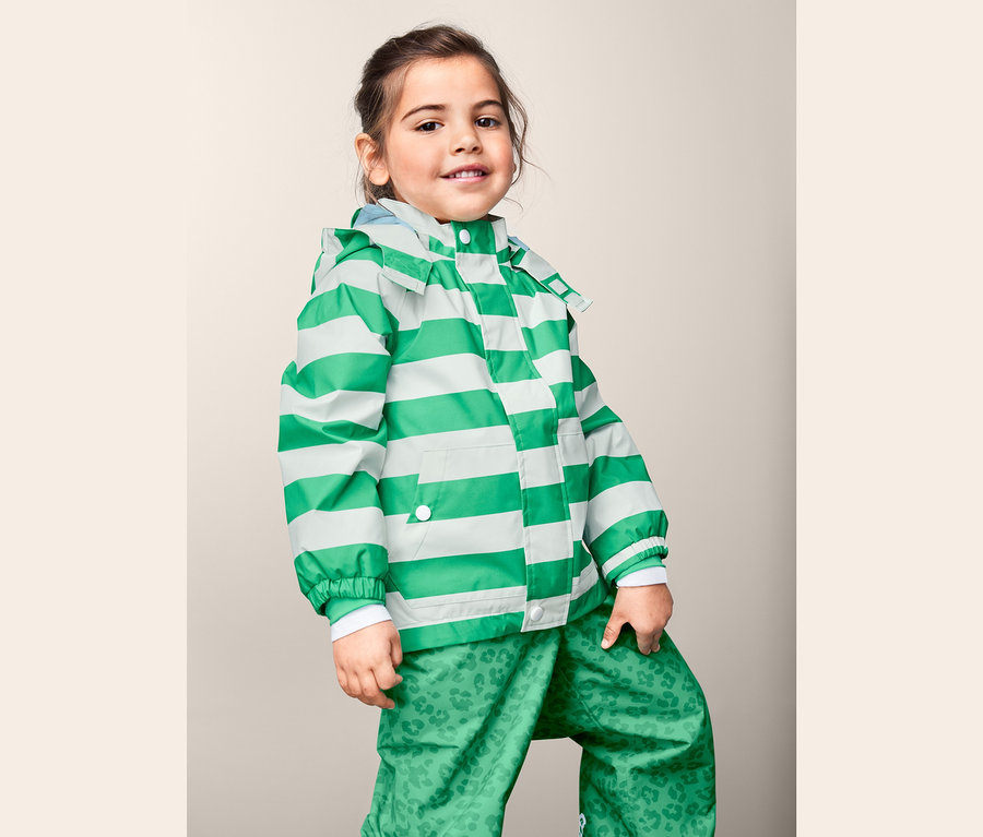 Kislány pózol egy zöld-fehér csíkos, újrahasznosított anyagból készült esőkabátban és egy zöld, mintás nadrágban.