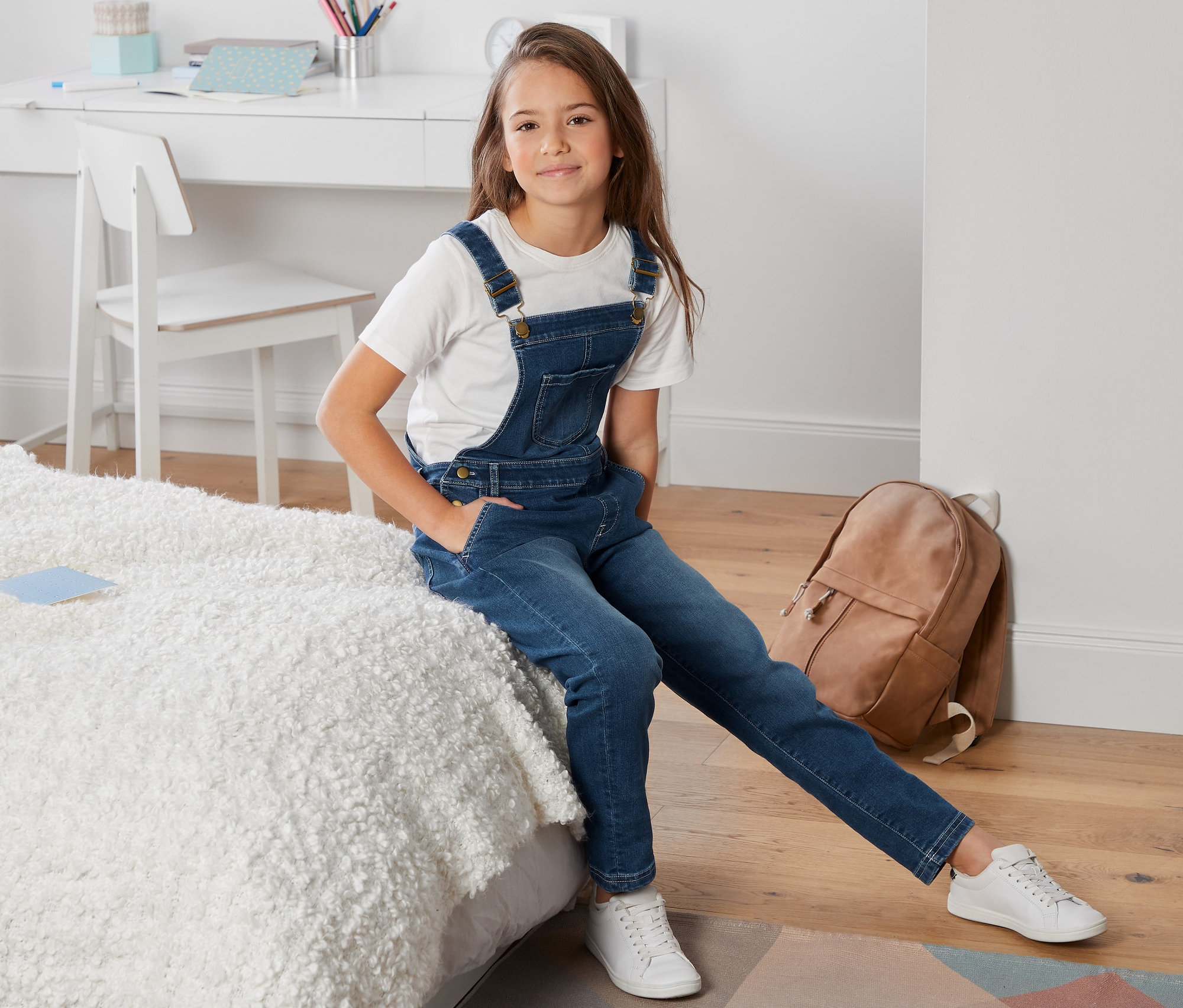 Egy lány ül az ágyon, kék kantáros farmernadrágot visel.