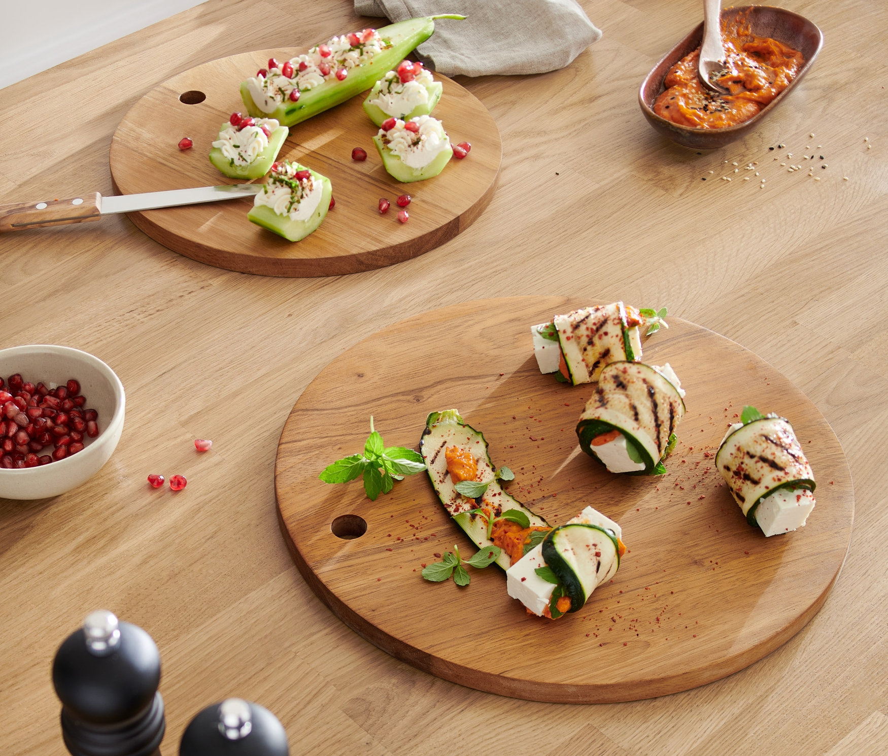 Két kerek vágódeszka cukkinitekercsekkel és töltött cukkinivel egy fa asztalon.