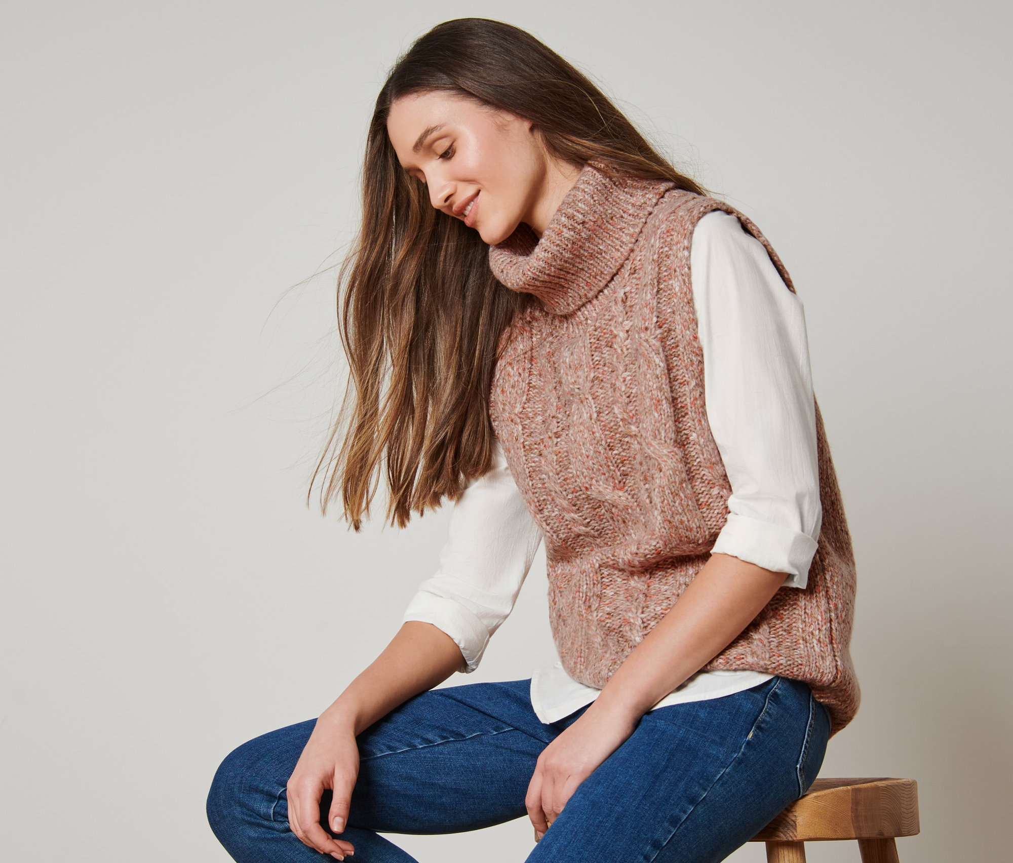 Nő ül egy széken, és kötött garbó mellényt visel.