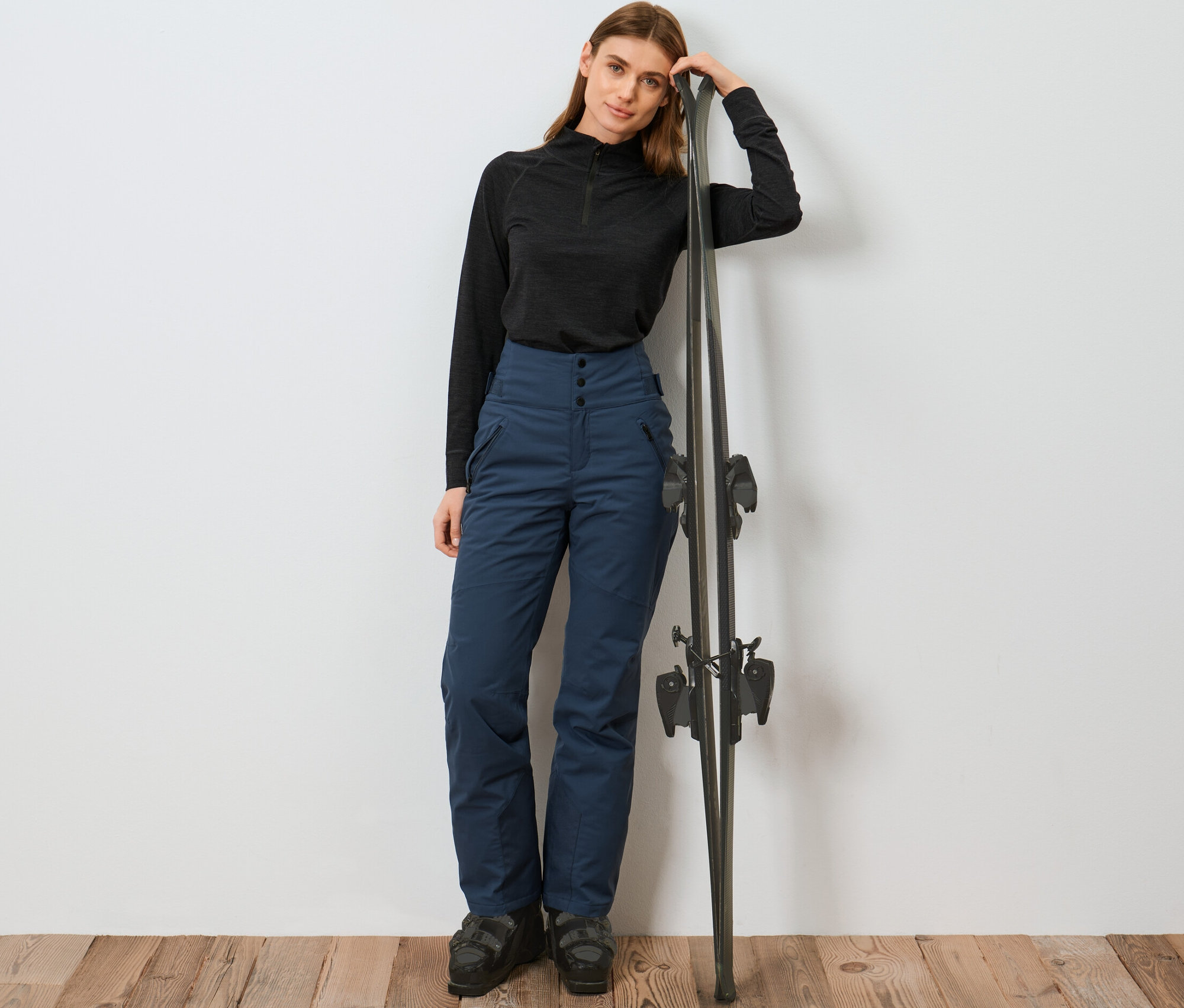 Egy nő a sílécnek támaszkodik, kék, magas derekú sínadrágot visel.