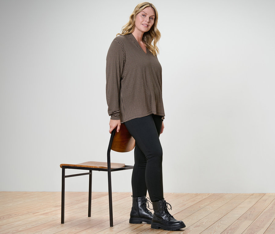 Nő pózol nyomott mintás pólóblúzban V-nyakkivágással és fekete leggingsben egy szék mellett.