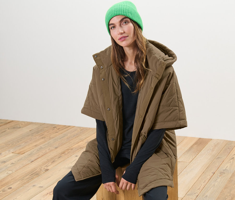 Nő ül egy fa ülőkén, barna steppelt poncsót és zöld sapkát visel.