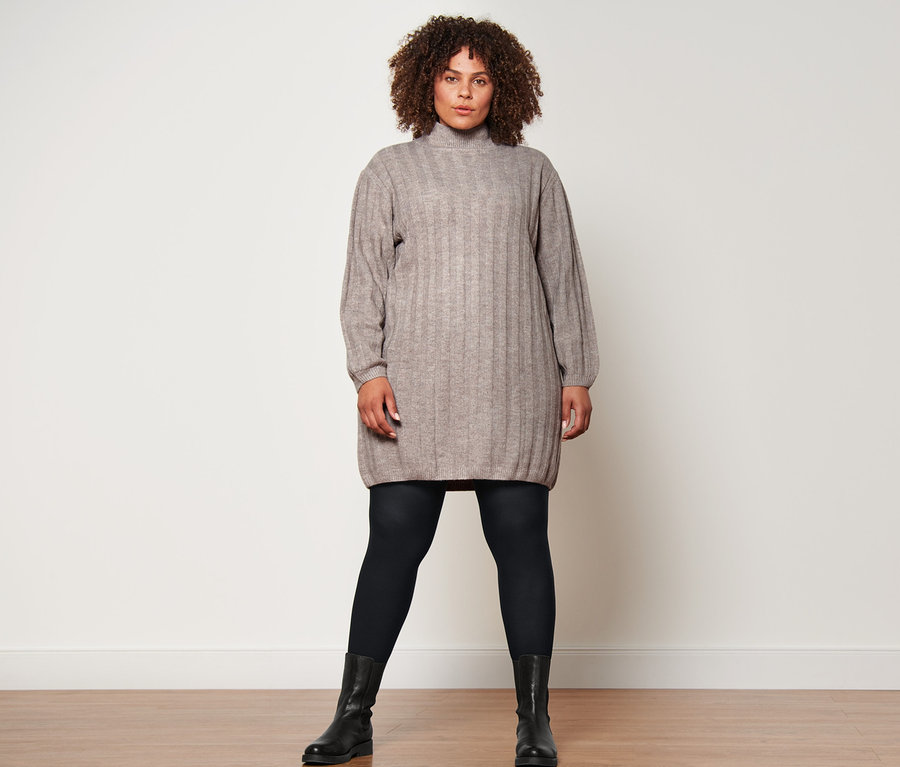 Egy nő áll kötött ruhában, fekete harisnyát és bőrcsizmát visel.