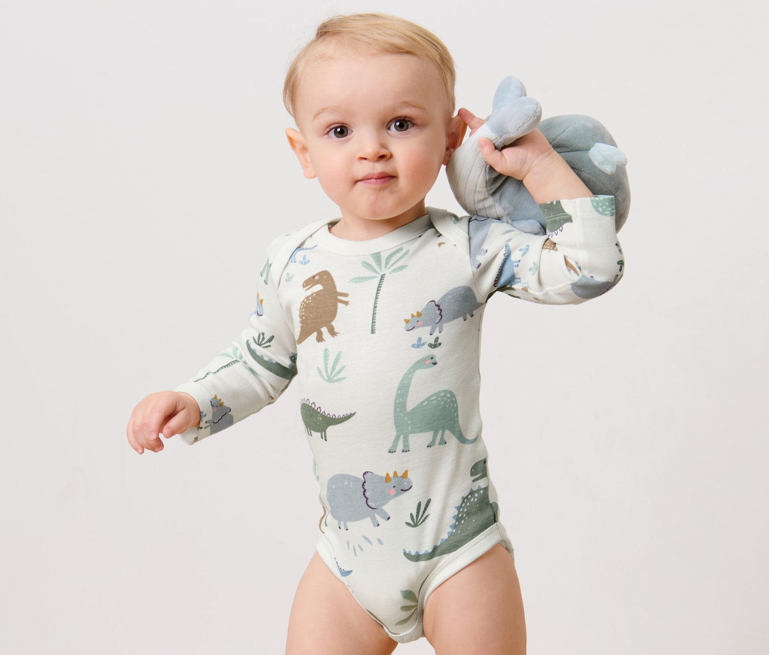 Egy baba plüssállatot tart és 3 hosszú ujjú body, dinó mintás bodyt visel.