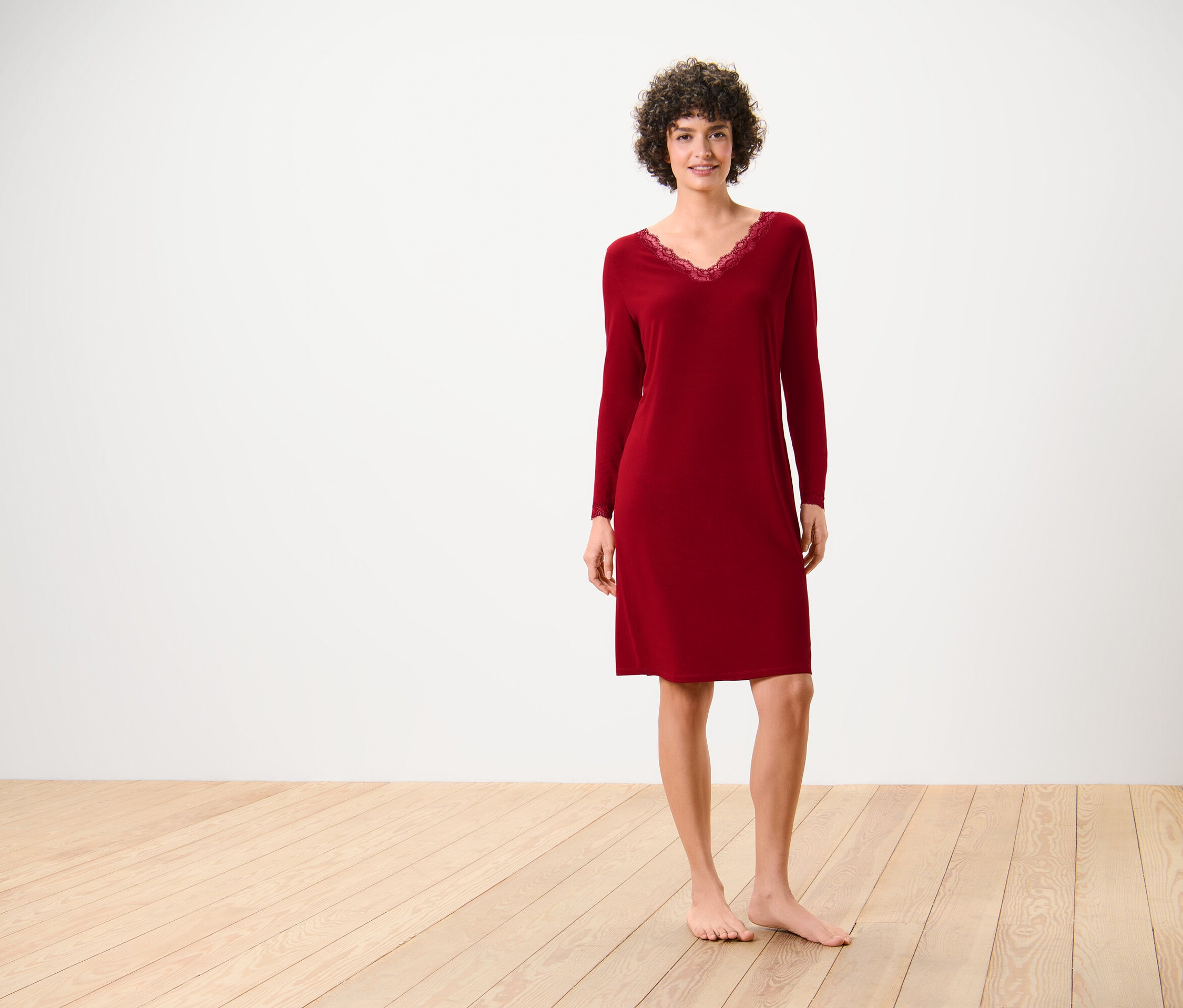 Egy nő vörös, hosszú ujjú, csipkés nyakú hálóingben áll egy fapadlón fehér fal előtt.