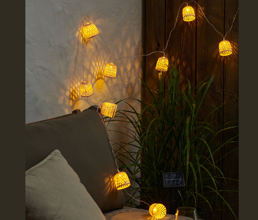Egy világító, dekoratív lámpabúrás napelemes fényfüzér lóg egy szürke kanapé felett.