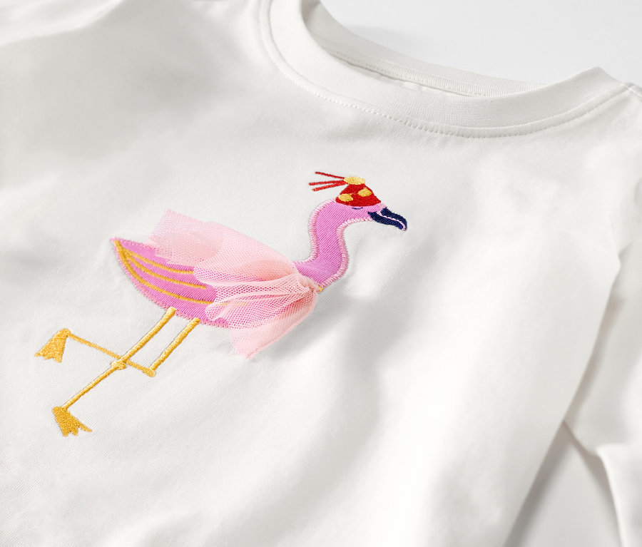 Egy hosszú ujjú gyerek póló részlete, hímzett flamingóval, tütüvel és születésnapi sapkával.