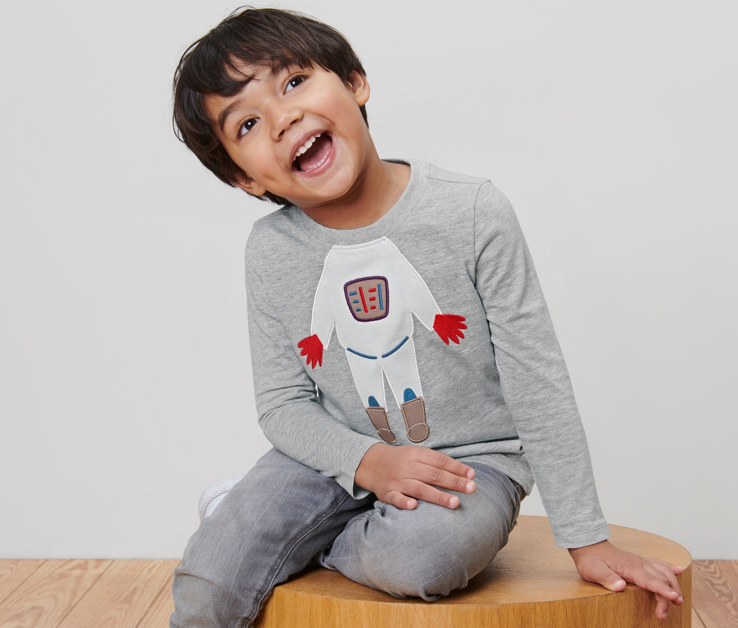 Egy fiú ül egy fa zsámolyon, és egy űrmintás, szürke, hosszú ujjú felsőt visel.