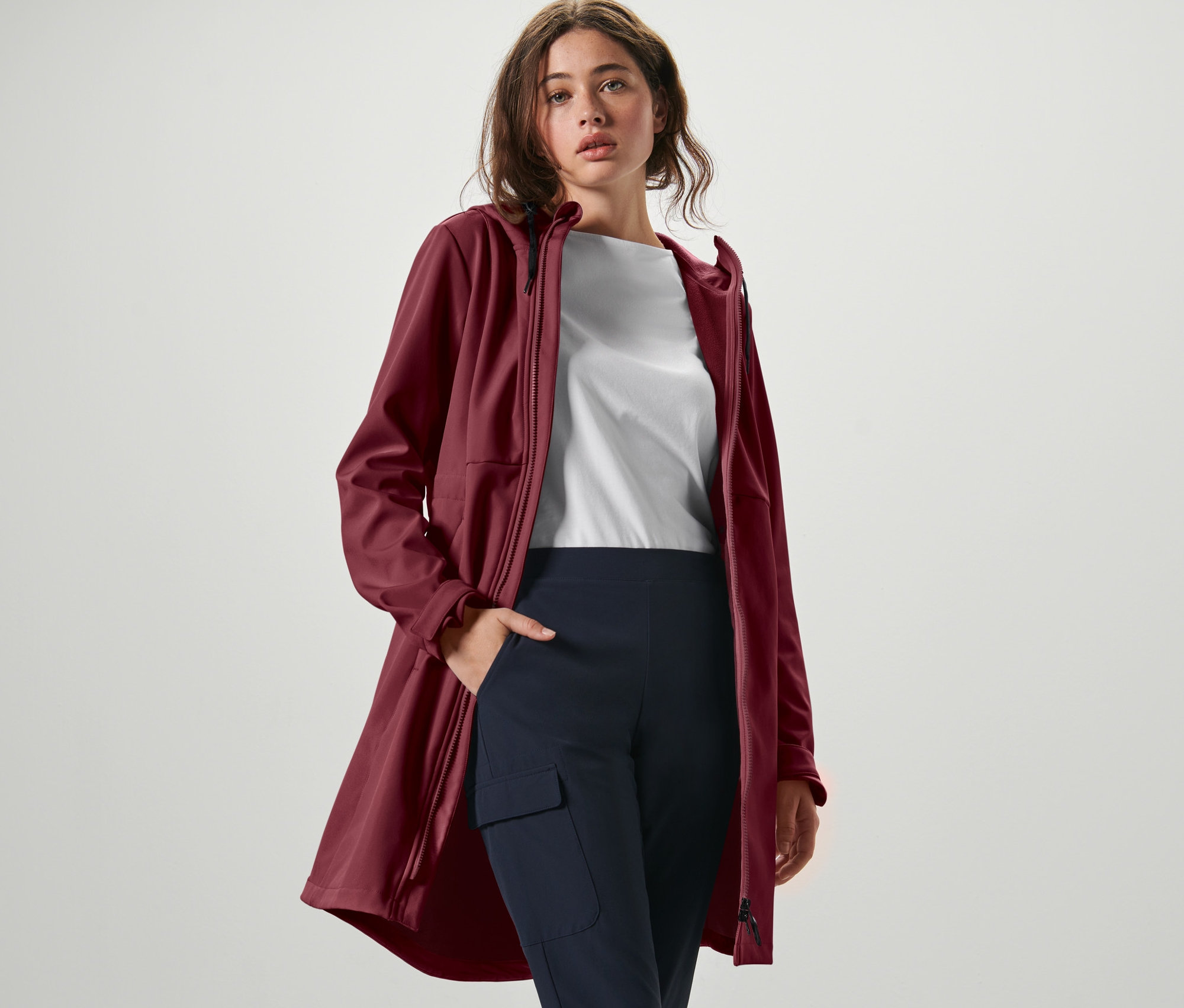 Nő pózol bordó softshell kabátban, fehér pólóban és sötétkék nadrágban.