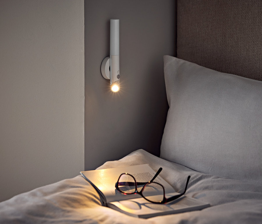 Egy ágyon szemüveg és egy nyitott könyv egy fali lámpa alatt.