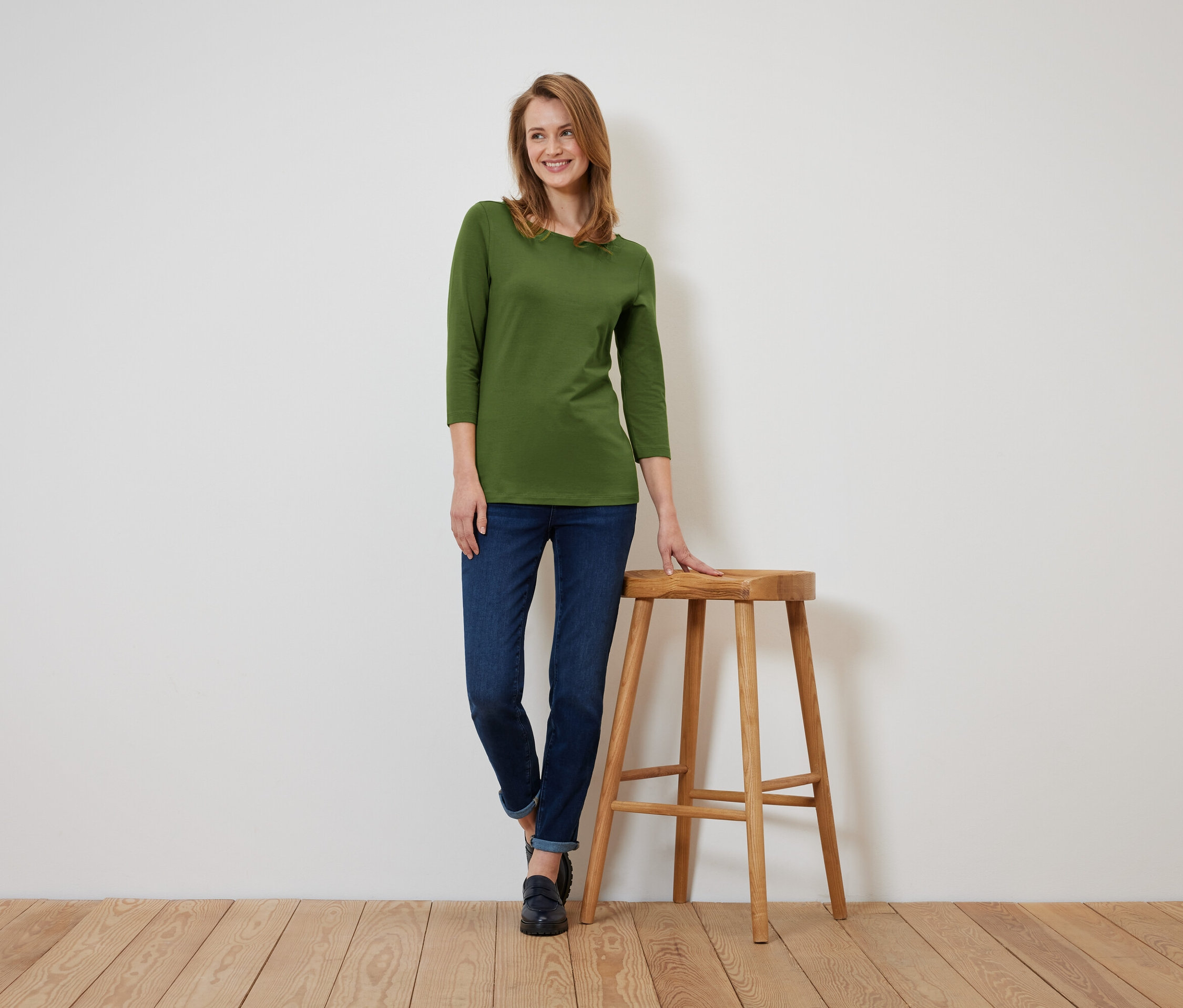 Egy olívazöld, háromnegyedes ujjú pólót és sötétkék farmert viselő nő áll és egy fa zsámolyra támaszkodik.