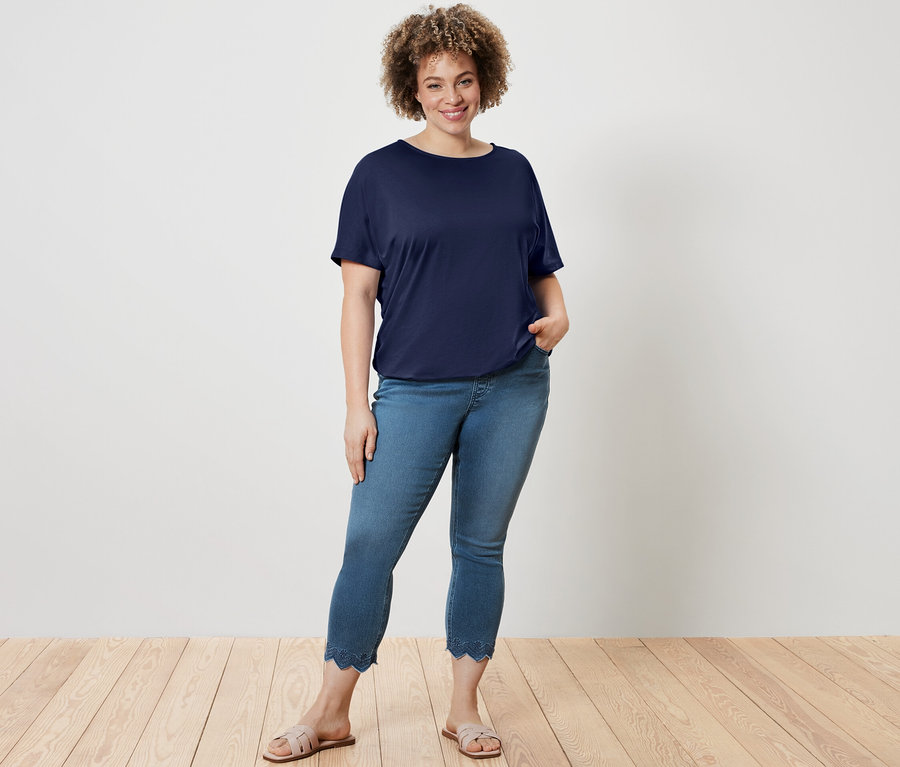 Nő pózol sötétkék női pólóblúzban és 7/8-os, lyukhímzéses treggingsben.