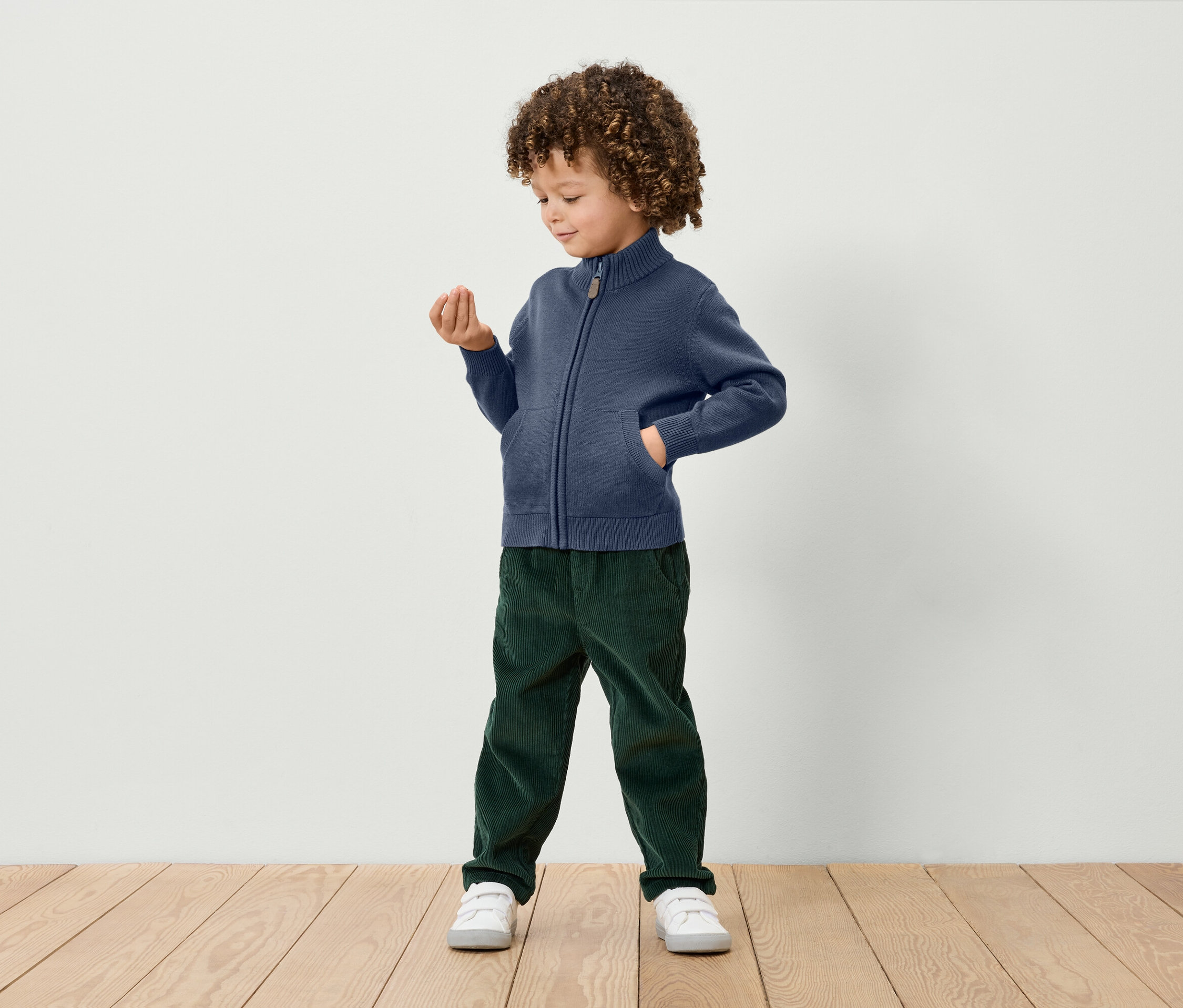 A göndör hajú fiú áll és egy kis tárgyat tart a kezében. Kék, cipzáras kötött kardigánt, zöld kordbársony nadrágot és fehér tornacipőt visel.