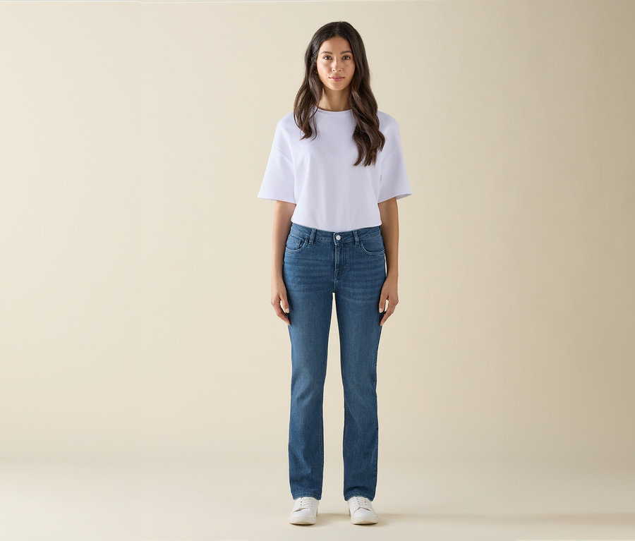 Egy nő áll, fehér pólót, kék farmert és fehér tornacipőt visel.