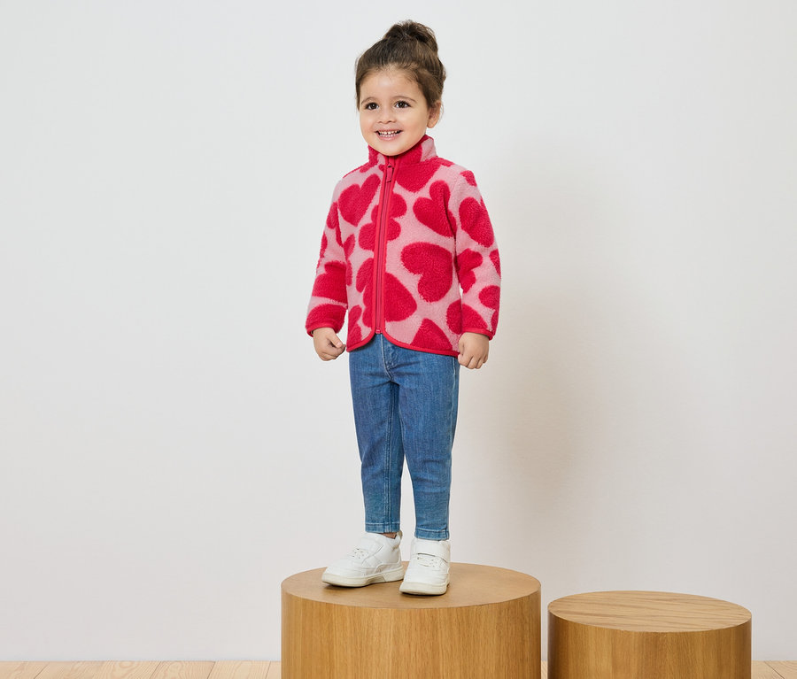 Mosolygó kislány áll egy fa talapzaton, rózsaszín, piros szíves mintás fleece kabátot, farmert és fehér tornacipőt visel.