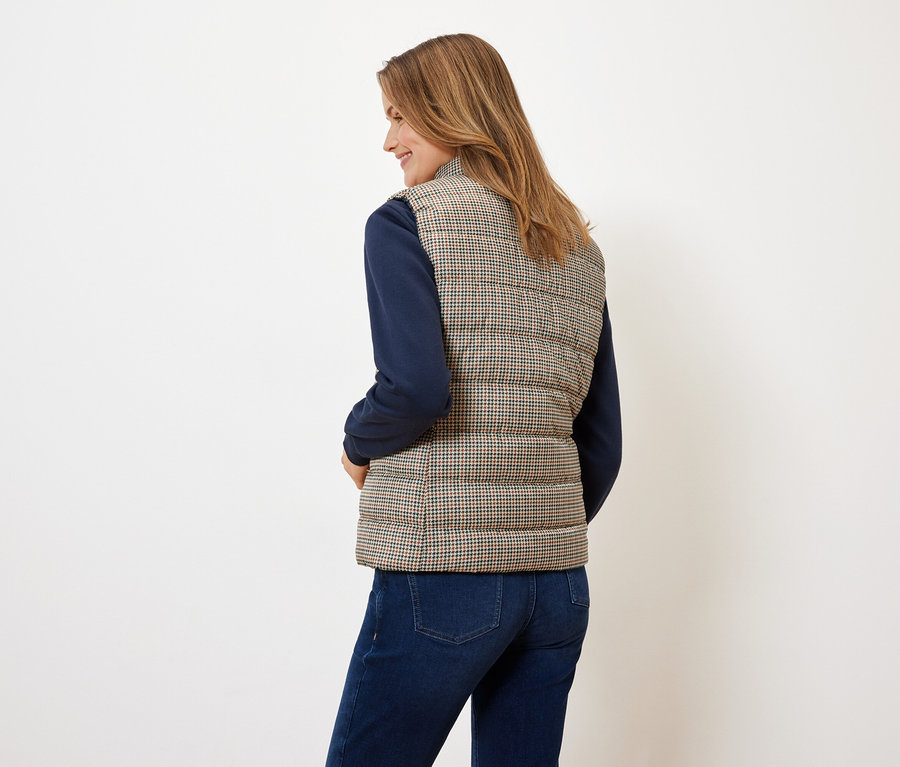 A fényképezőgépnek háttal álló nő tyúkláb mintás steppelt mellényt, kék pulóvert és farmert visel.