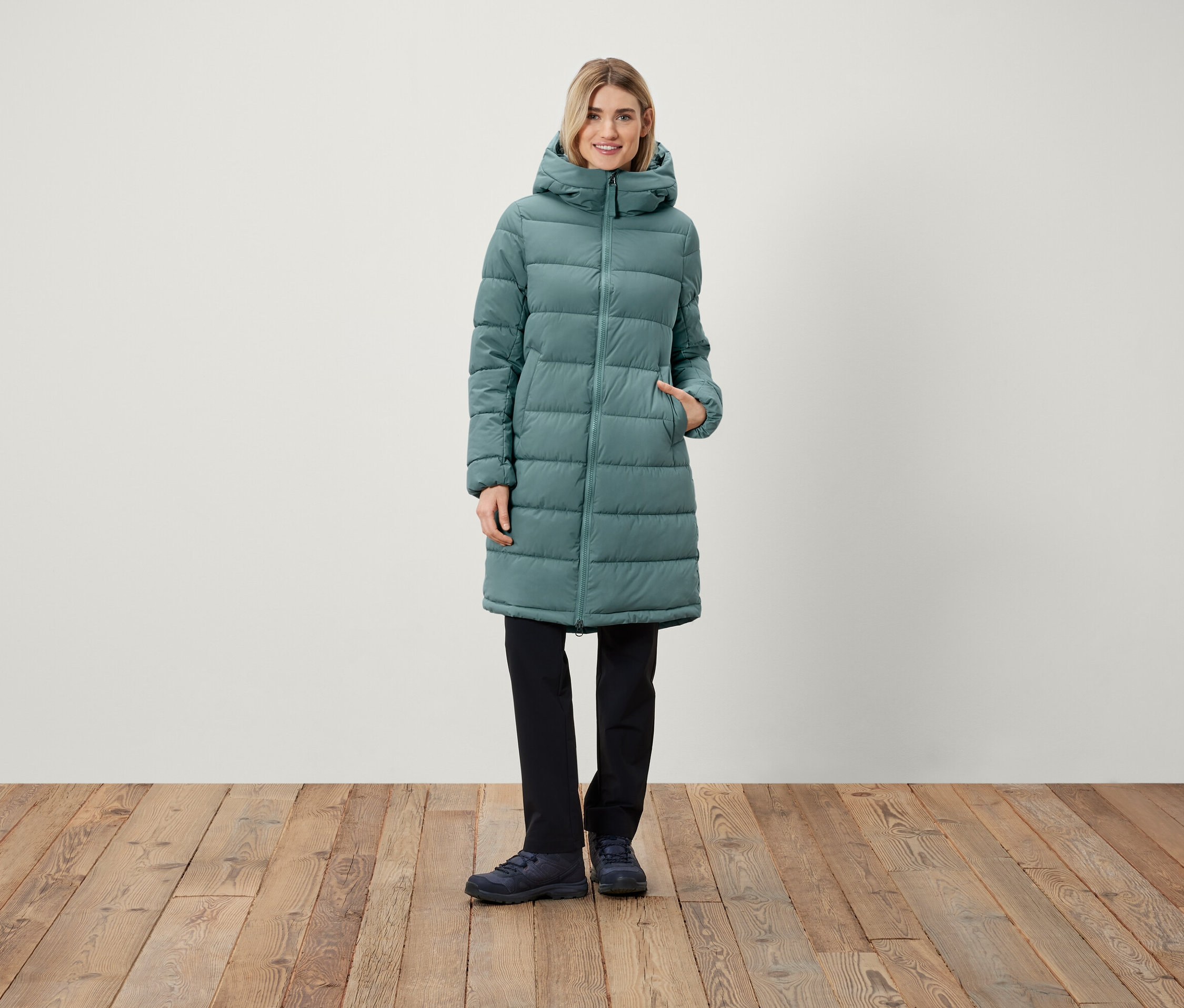 Egy nő áll, hosszú, kapucnis steppelt kabátot, fekete nadrágot és cipőt visel.