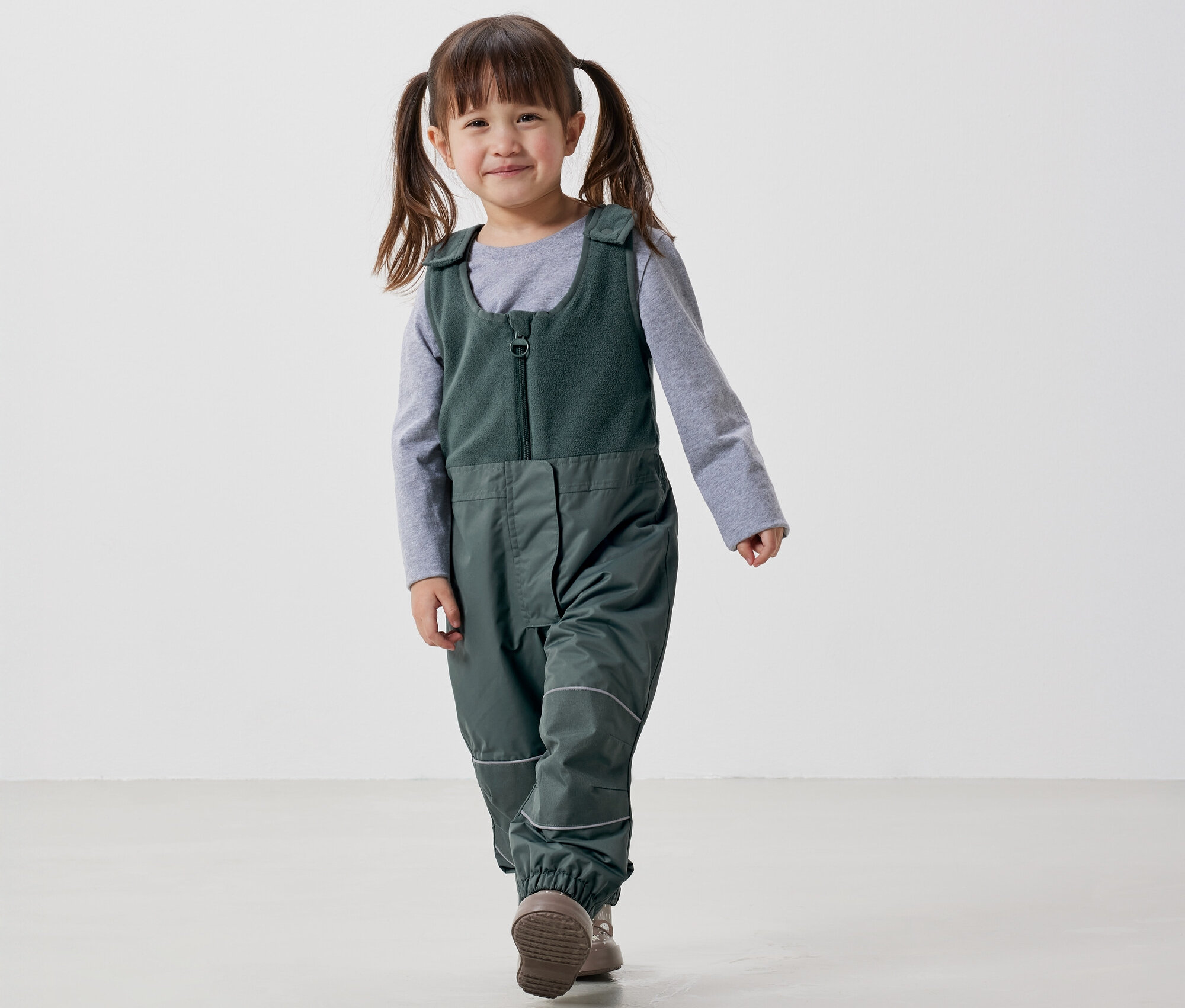 Mosolygó kislány copfokkal zöld kantáros nadrágban és szürke pólóban sétál fehér háttér előtt.