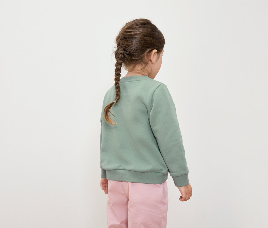 Hátulnézet egy zöld pulóvert és rózsaszín nadrágot viselő, fonott hajú lányról.