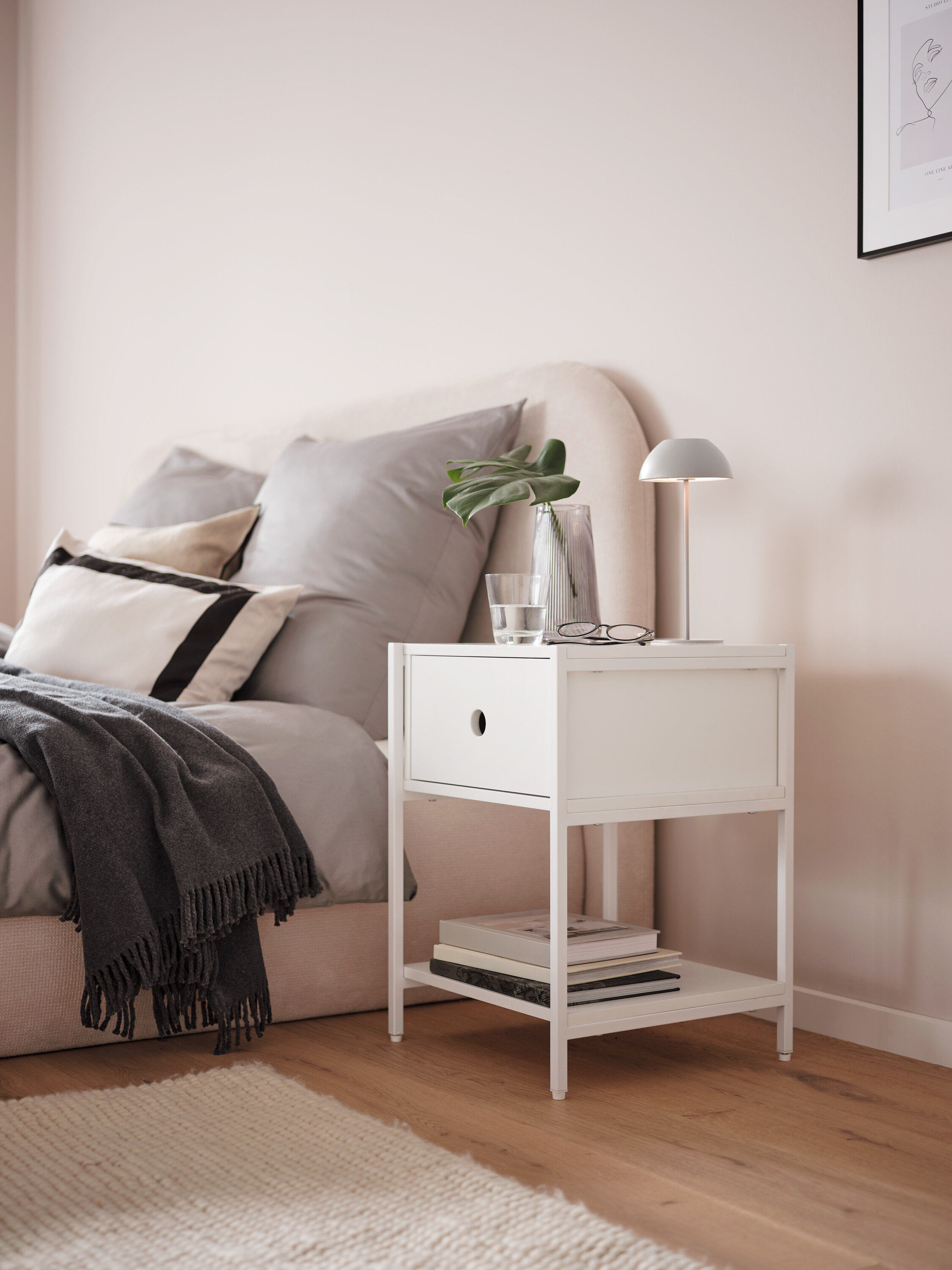 Szürke LED-es akkus asztali lámpa egy fehér éjjeliszekrényen, egy ágy mellett.