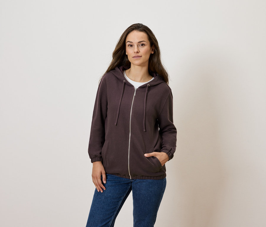 Nő pózol barna, cipzáras pulóverben és kék farmerben.