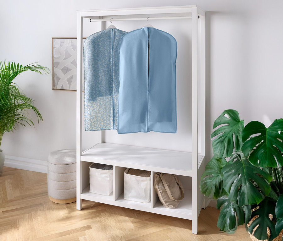Két ruhazsák lóg egy fehér szekrényen, alatta polcokkal és rekeszekkel. Balra és jobbra egy-egy szobanövény áll.