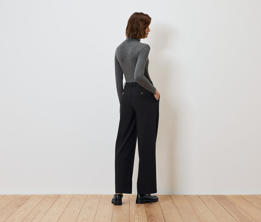 Hátulról látható egy nő, kezével a fekete nadrágja zsebében, szürke garbóban.