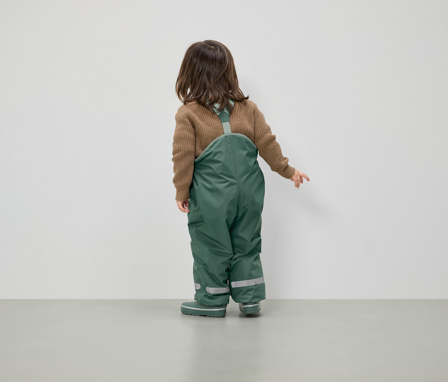 Egy barna pulóvert és zöld kantáros nadrágot viselő gyermek háttal áll a kamerának.