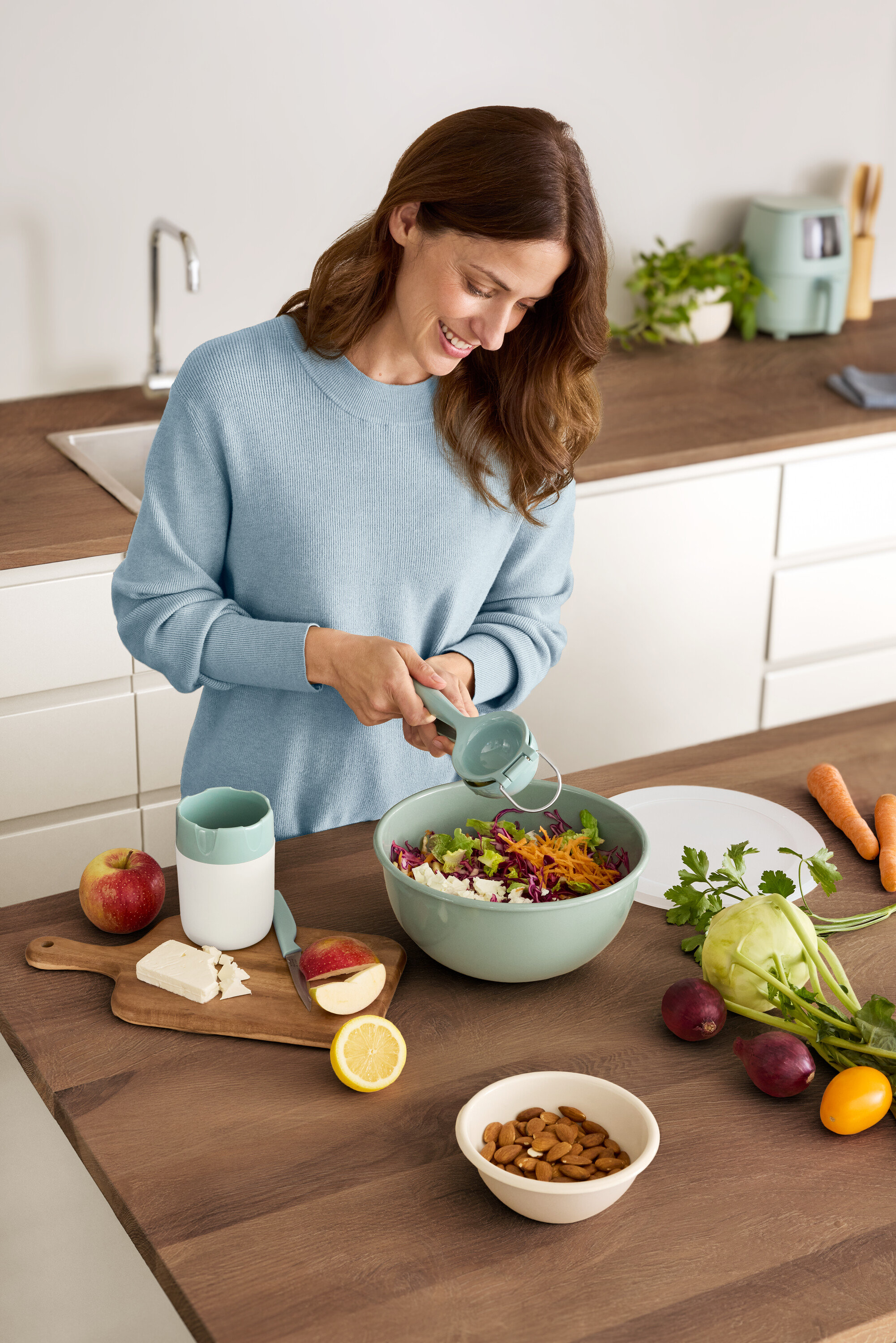 Nő citromot facsar egy salátástálba. Az asztalon alma, sajt, mandula és zöldségek találhatók.