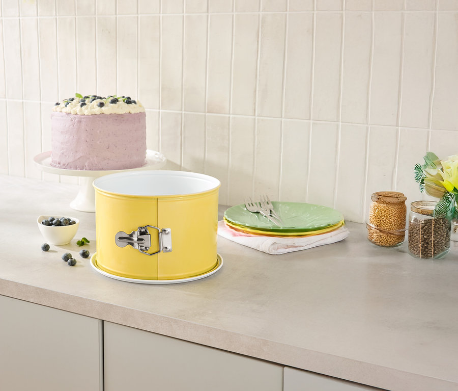 A konyhapulton egy sárga tortaforma, egy torta tortatálon, tányérok és tárolóedények állnak.