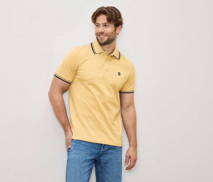 Férfi sárga pólóingben és farmerban áll egy világos fal előtt.