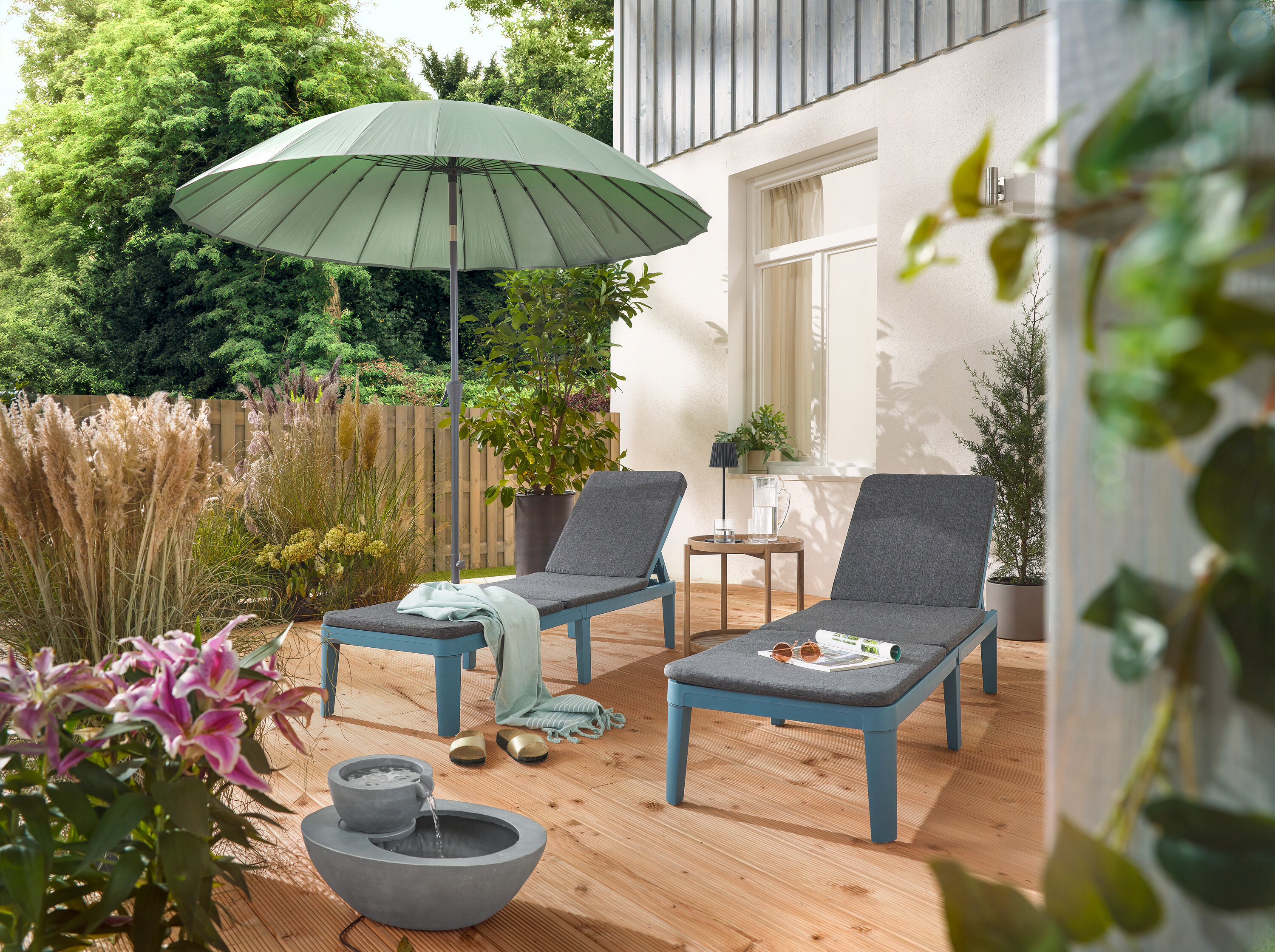 A teraszon két nyugágy áll egy világosszürke Leco Shanghai napernyő alatt és egy napelemes kerti csobogó mellett.