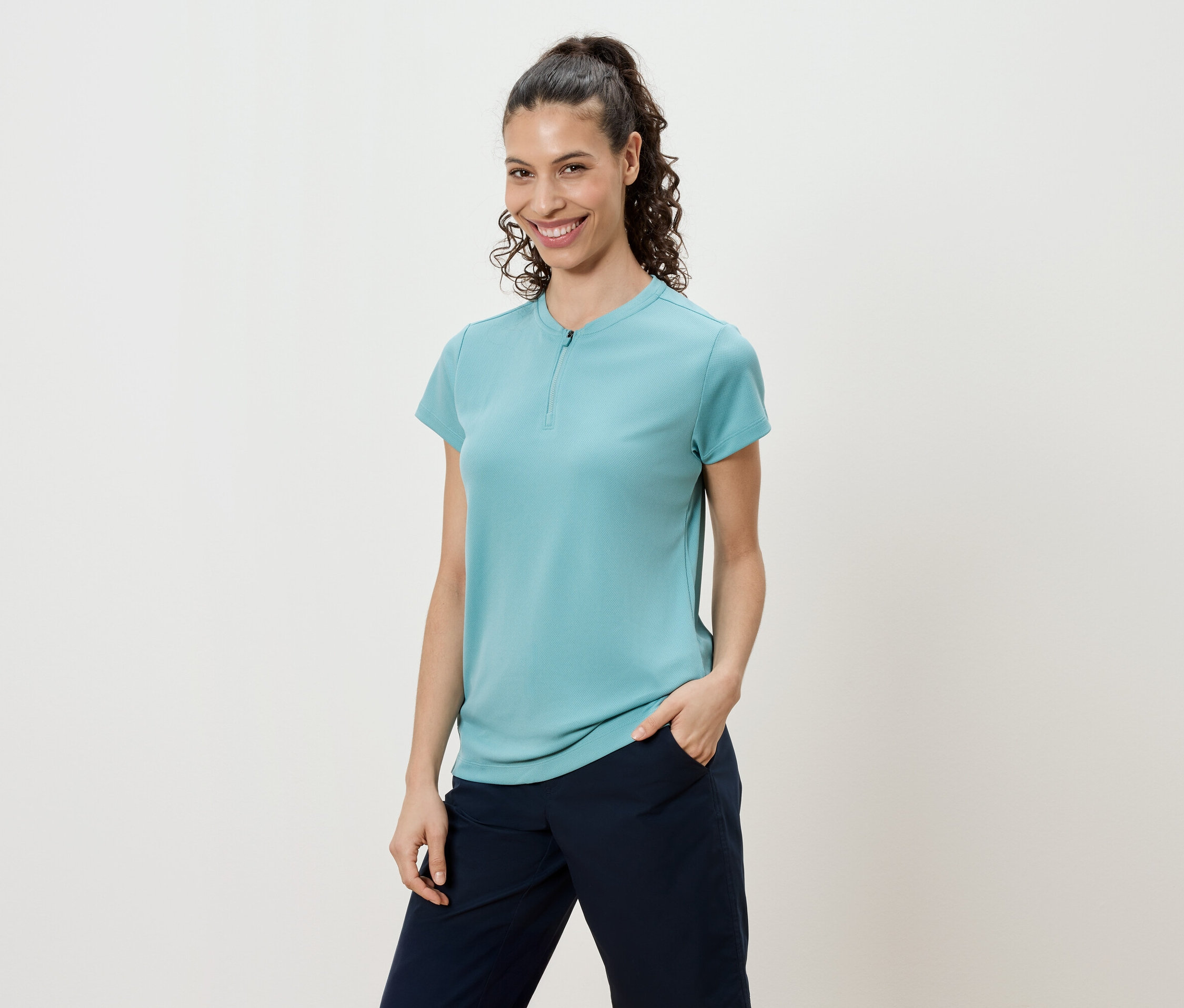 Lófarokba kötött, göndör hajú nő türkizkék pólót és sötétkék nadrágot visel.