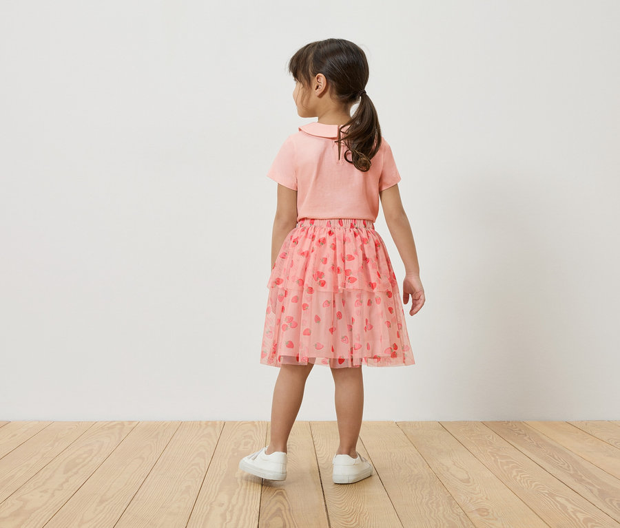 Egy kislány háttal áll a kamerának, rózsaszín pólót, rózsaszín tüllszoknyát visel epermintával és fehér tornacipőt.