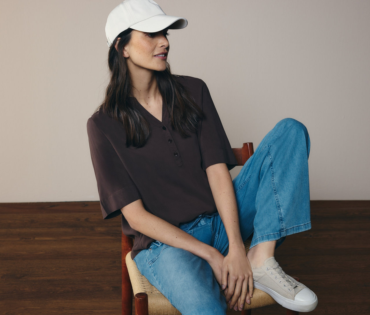 Nő ül a széken, farmert, sapkát és barna CECIL tunikablúzt visel.