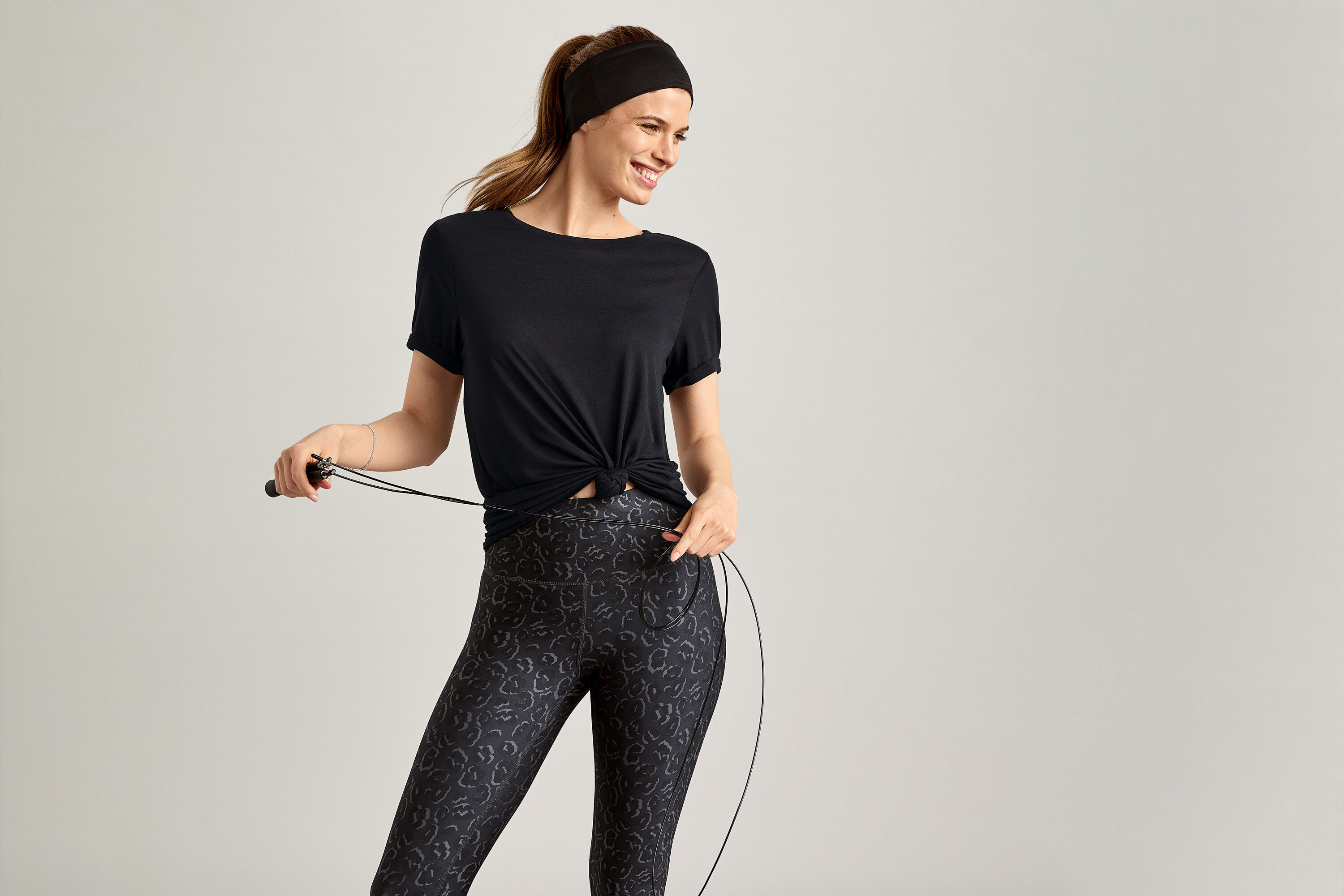 Nő fekete sportfelsőben ugrókötelet tart szürke háttér előtt.