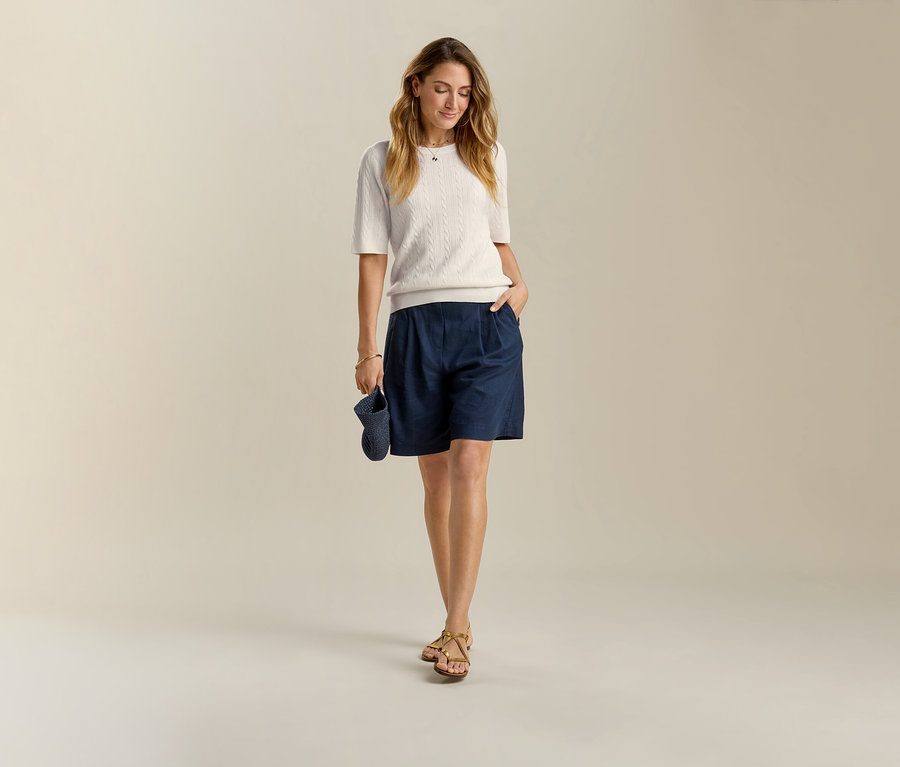 Nő sötétkék Leinen-Bermuda rövidnadrágban és szandálban. A kezében sötétkék sapkát tart.