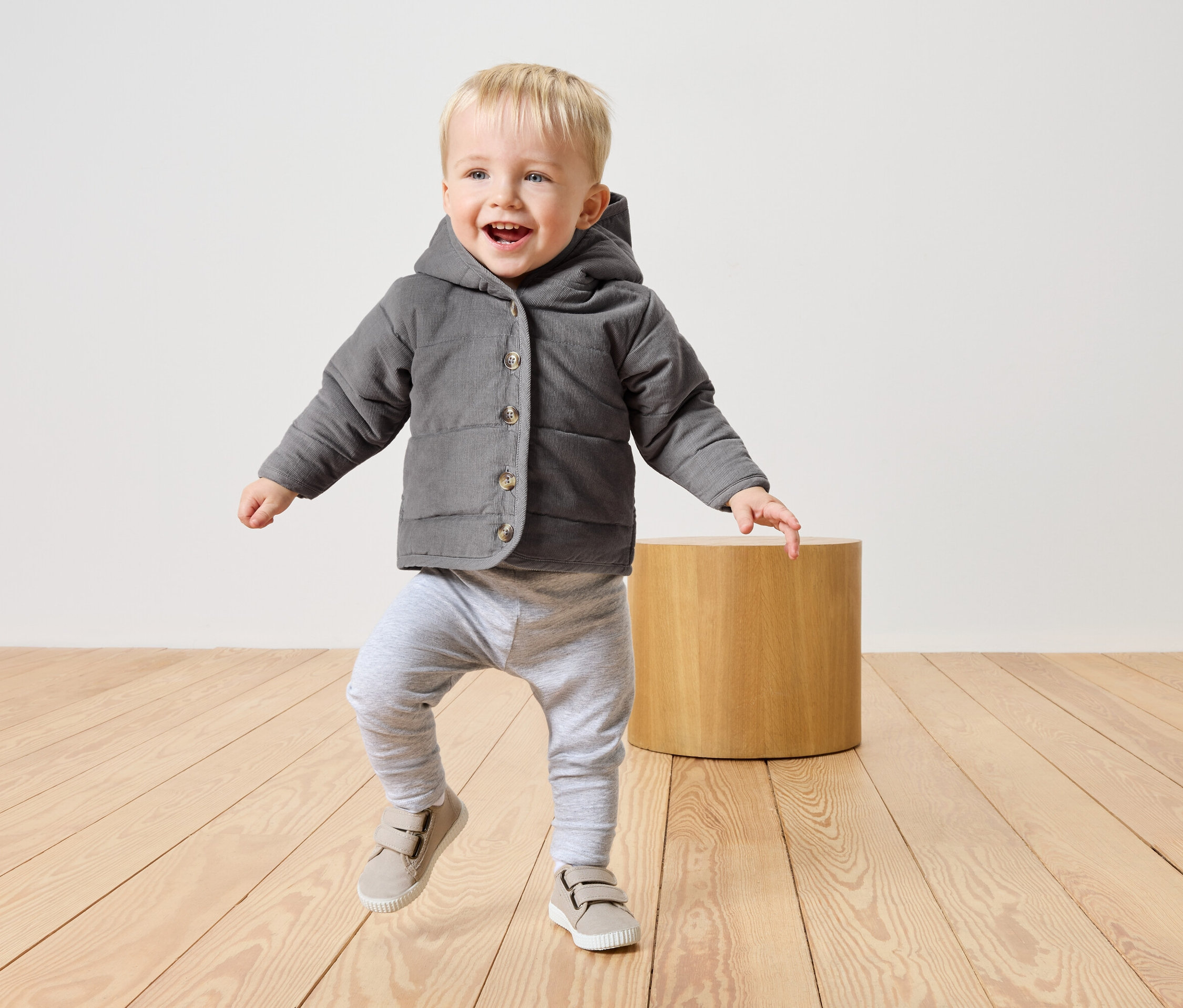 Egy kis szőke hajú fiú szürke kapucnis kabátban, szürke nadrágban és bézs cipőben sétál egy világos fa padlón.