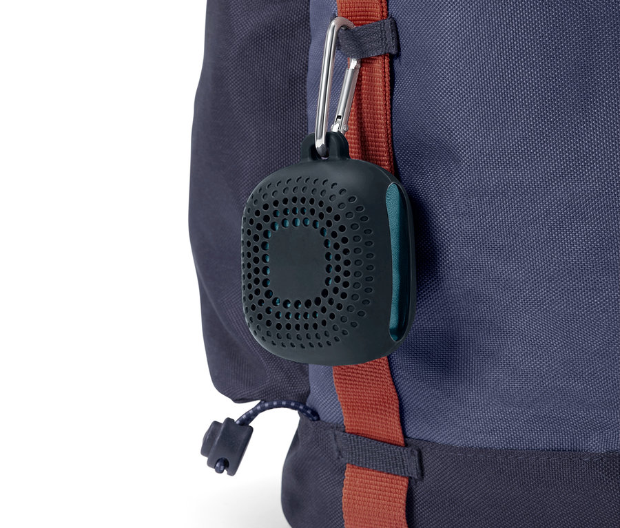 Egy fekete, kerek, lyukacsos tok karabinerrel van rögzítve egy kék hátizsákra.