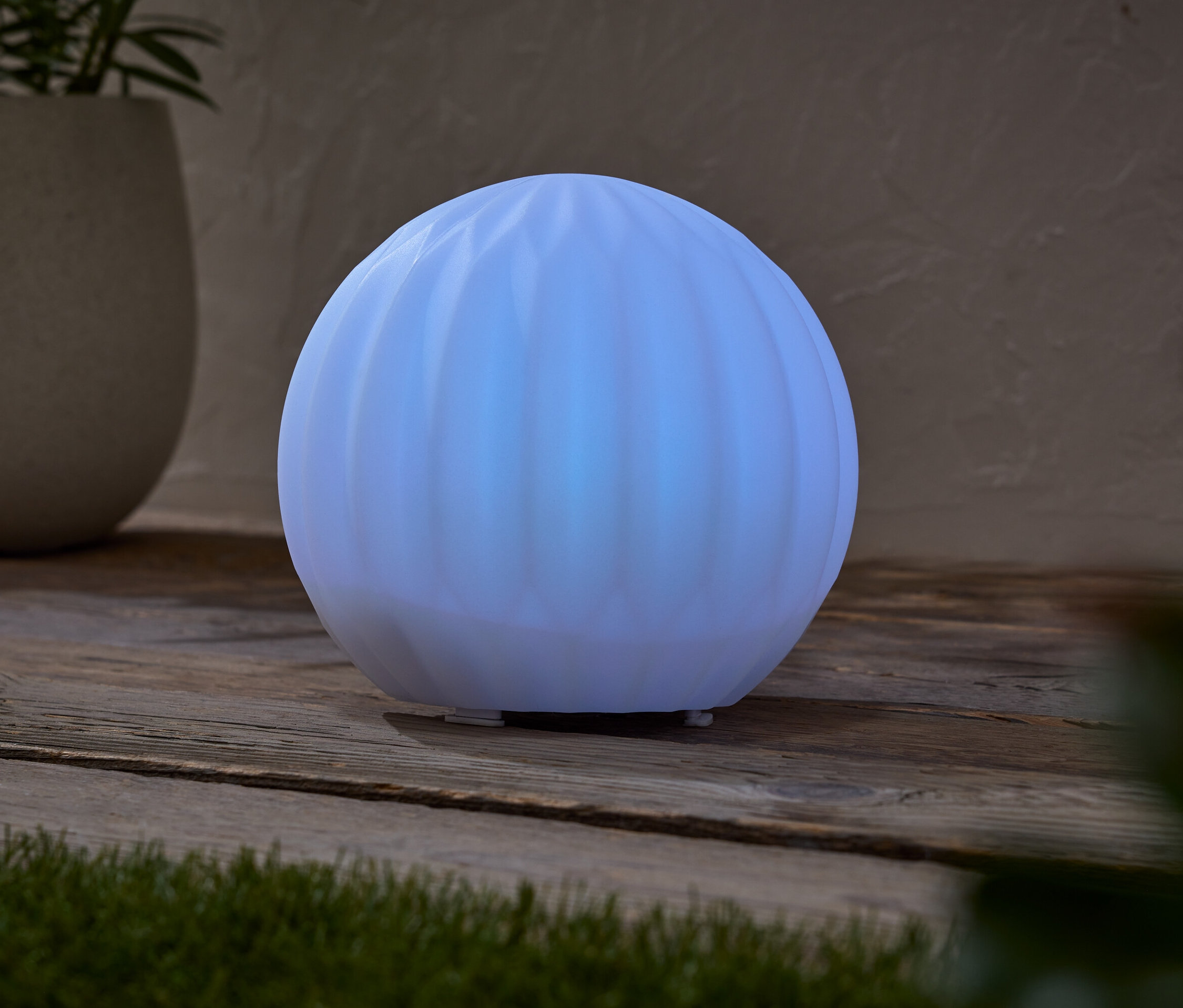 Egy világító, gömb alakú lámpa áll egy faasztalon, egy cserepes növény mellett.