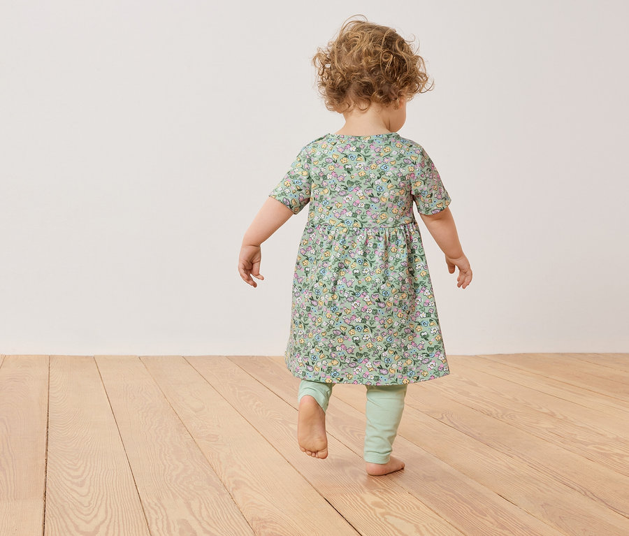 Egy kisgyermek göndör hajjal mezítláb sétál egy zöld, virágos ruhában és leggingsben egy fapadlón.