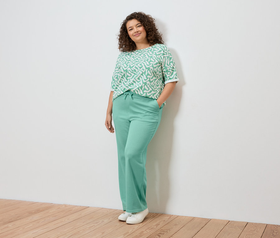 Nő pózol a falnál, világoszöld mintás felsőt, világoszöld nadrágot és fehér tornacipőt visel.