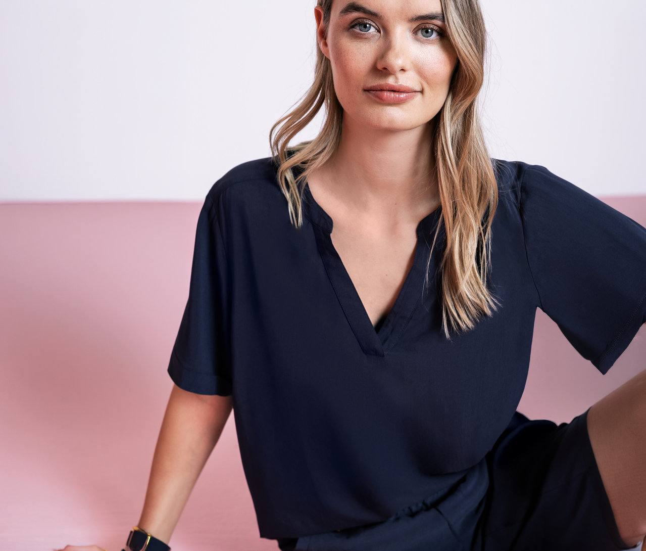 Nő ül rózsaszín háttér előtt, sötétkék Street One blúzban.